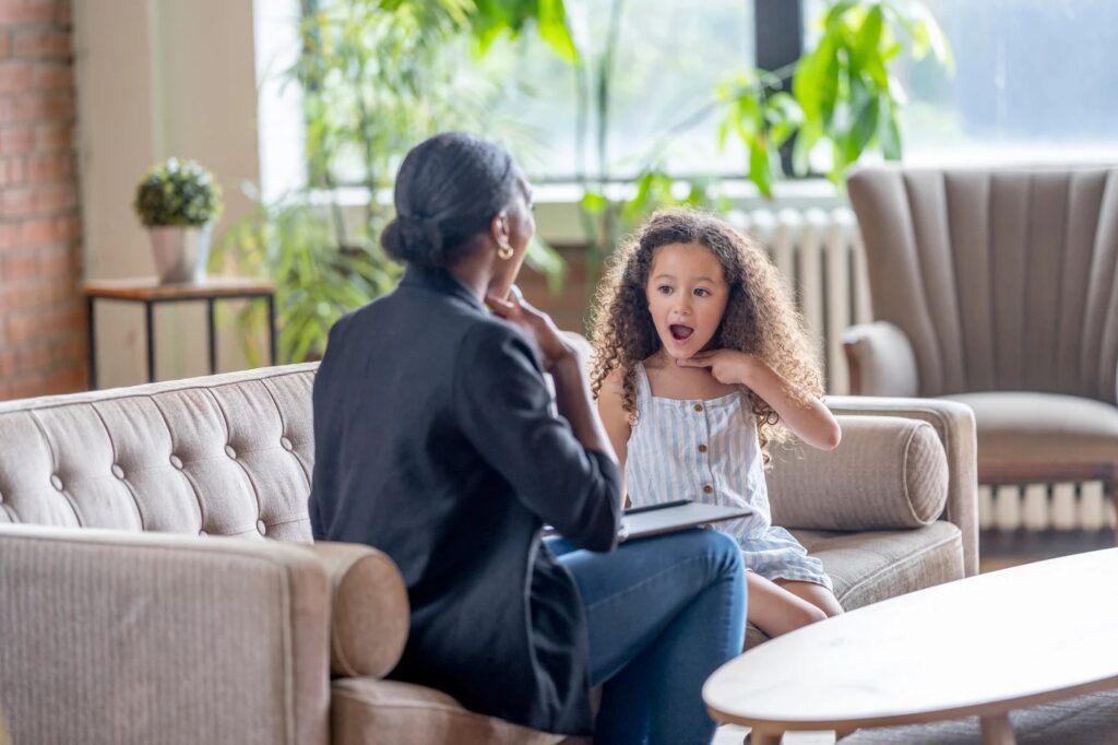Young child practicing speech therapy with speech language pathologist cueing for sound and modeling while sitting on a couch in front of a coffee table